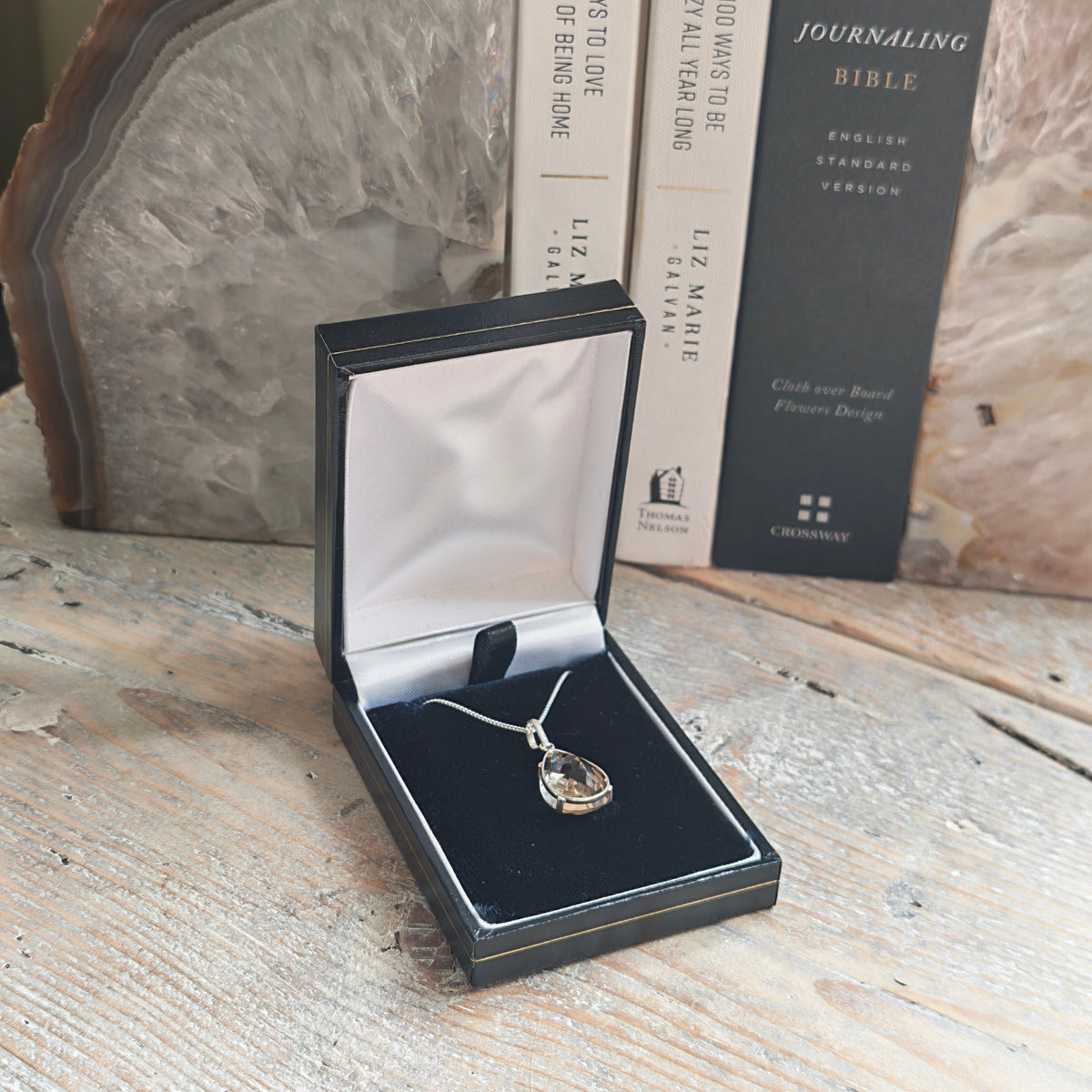 A silver pendant necklace with a teardrop-shaped citrine stone displayed in an open black presentation box on a rustic wooden surface, with books and an agate slice in the background.