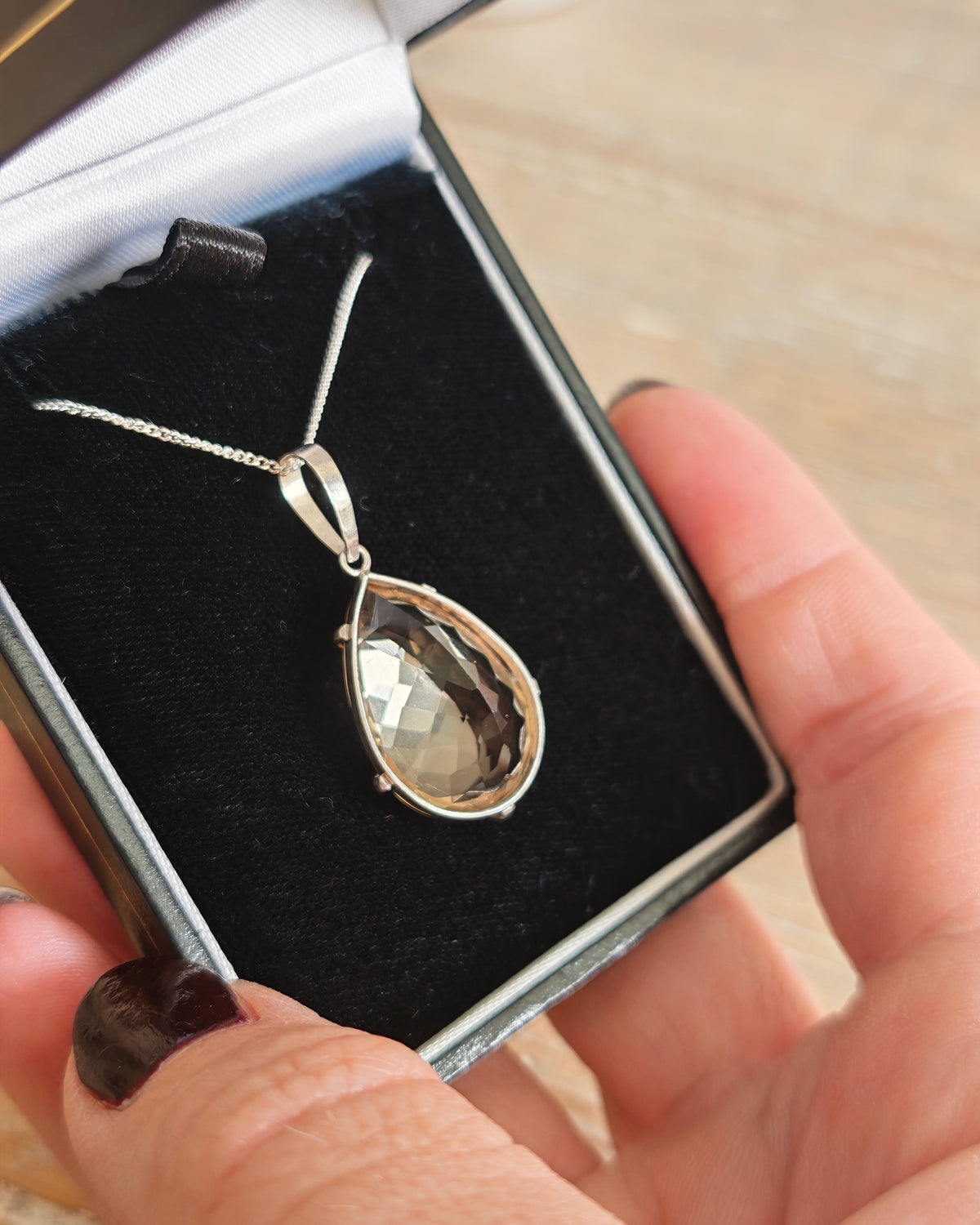 A citrine hand faceted stone in a silver teardrop pendant necklace , presented in a black velvet jewelry box held by a hand.