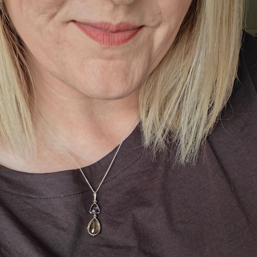 A silver pendant necklace featuring a larger citrine teardrop and a smaller triangular amethyst gemstone, being worn