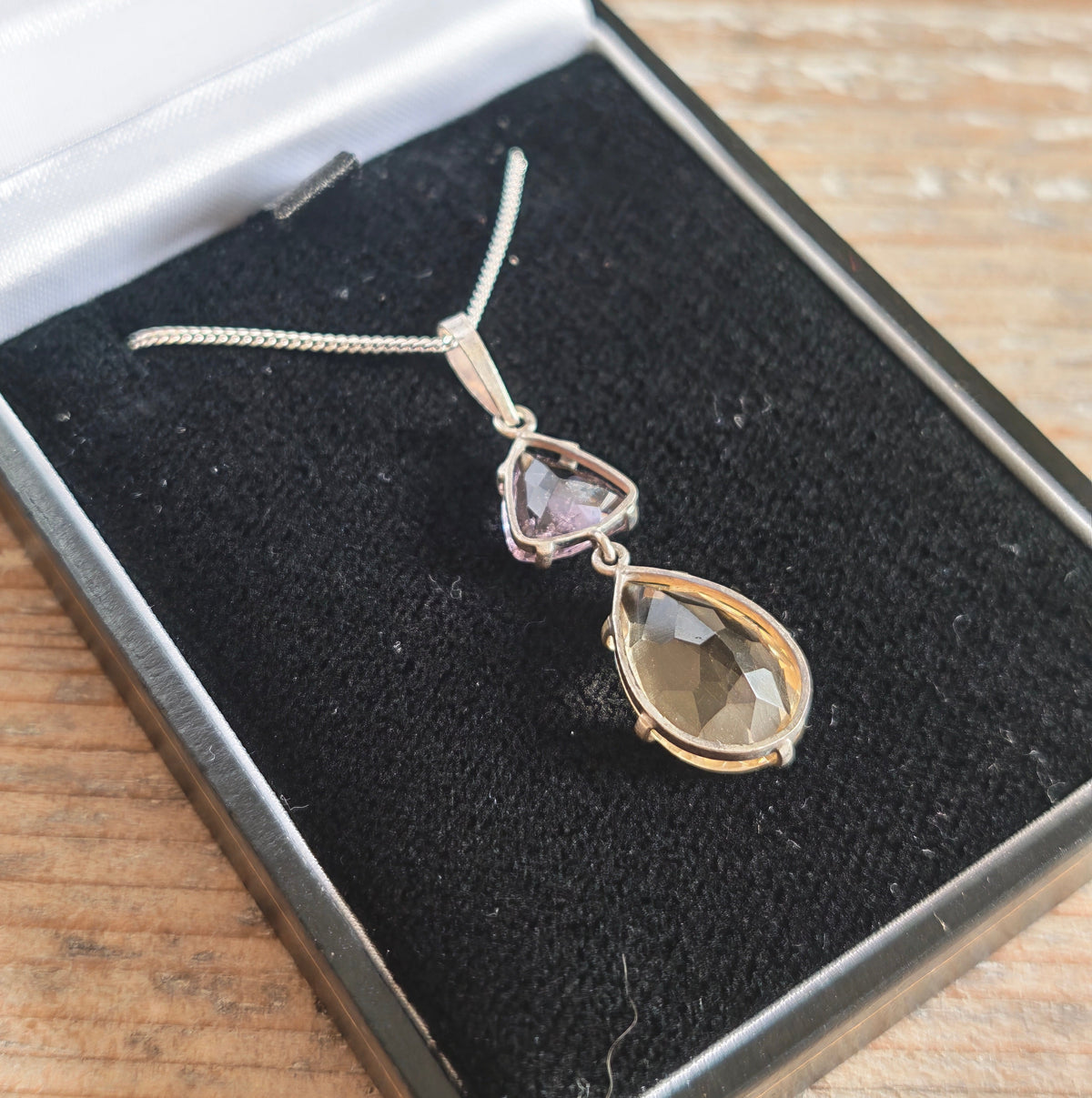 A silver pendant necklace featuring a larger citrine teardrop and a smaller triangular amethyst gemstone, in box on wood table closeup