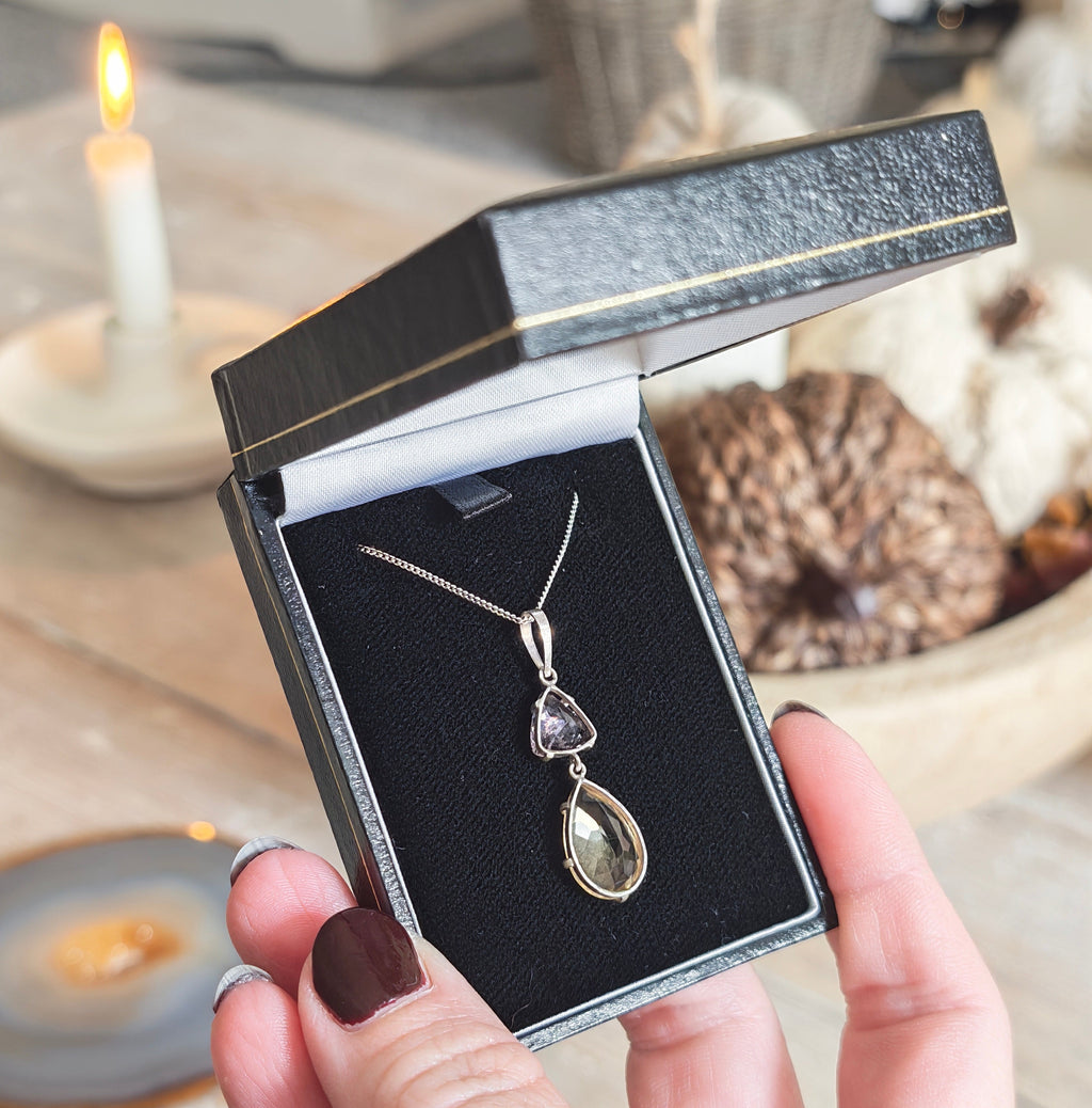 A silver pendant necklace featuring a larger citrine teardrop and a smaller triangular amethyst gemstone, in box held up