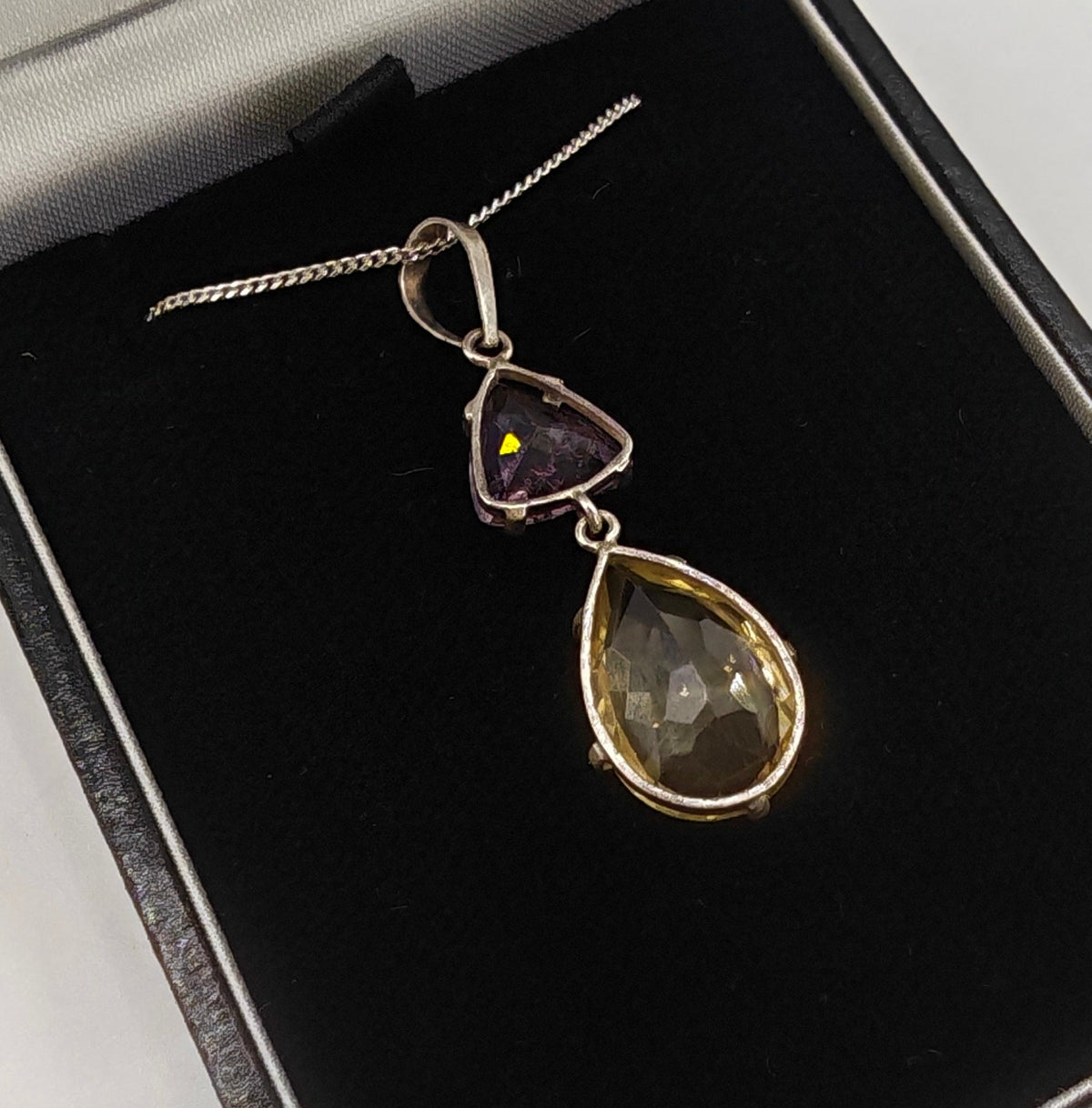 A silver pendant necklace featuring a larger citrine teardrop and a smaller triangular amethyst gemstone, in box close up