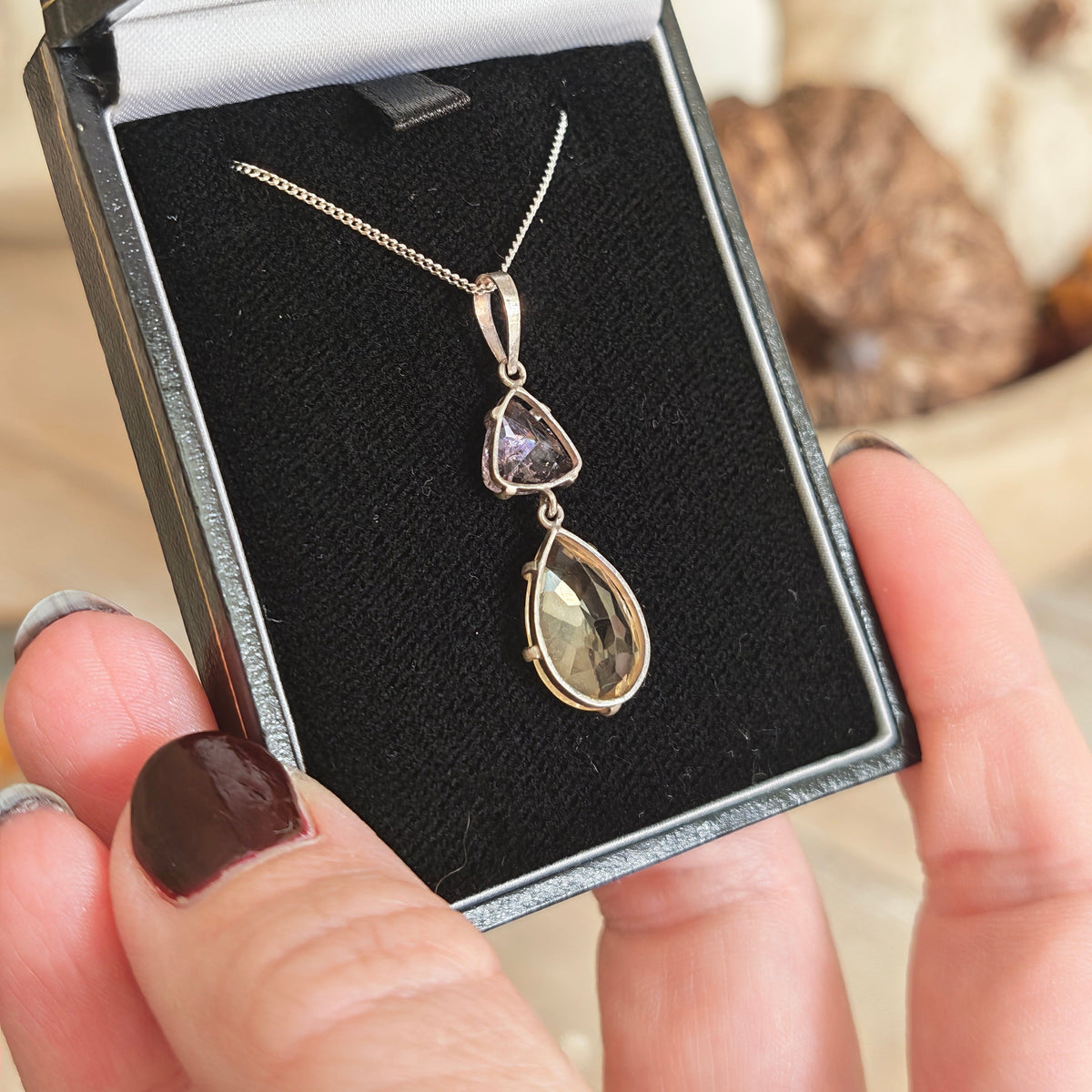 A silver pendant necklace featuring a larger citrine teardrop and a smaller triangular amethyst gemstone, in box held up closeup
