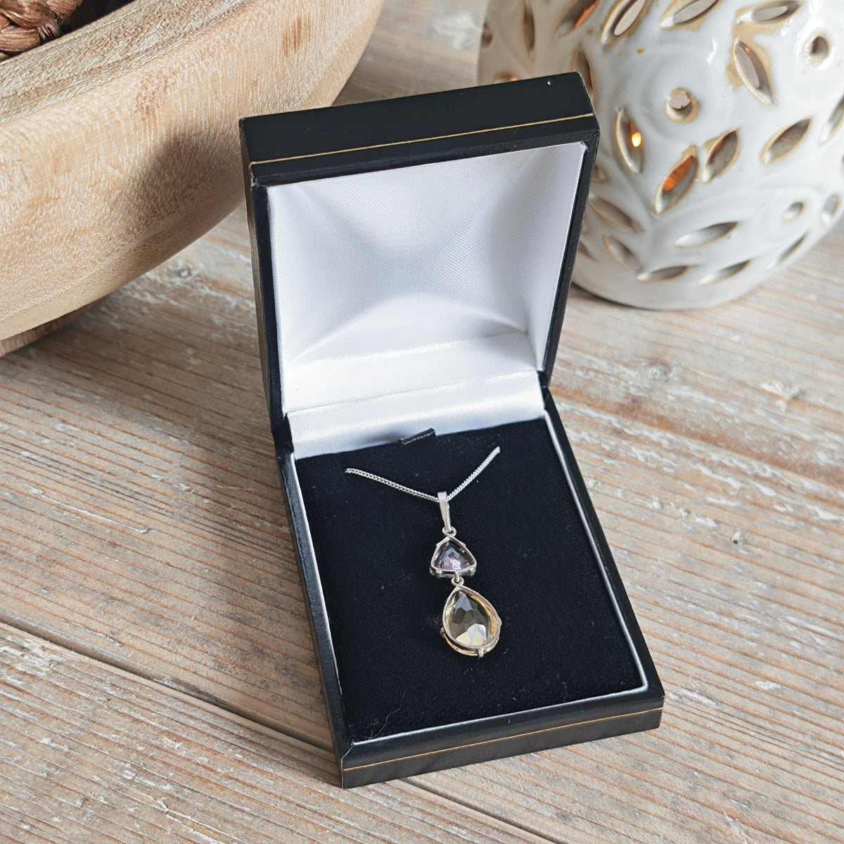 A silver pendant necklace featuring a larger citrine teardrop and a smaller triangular amethyst gemstone, in box on wood table