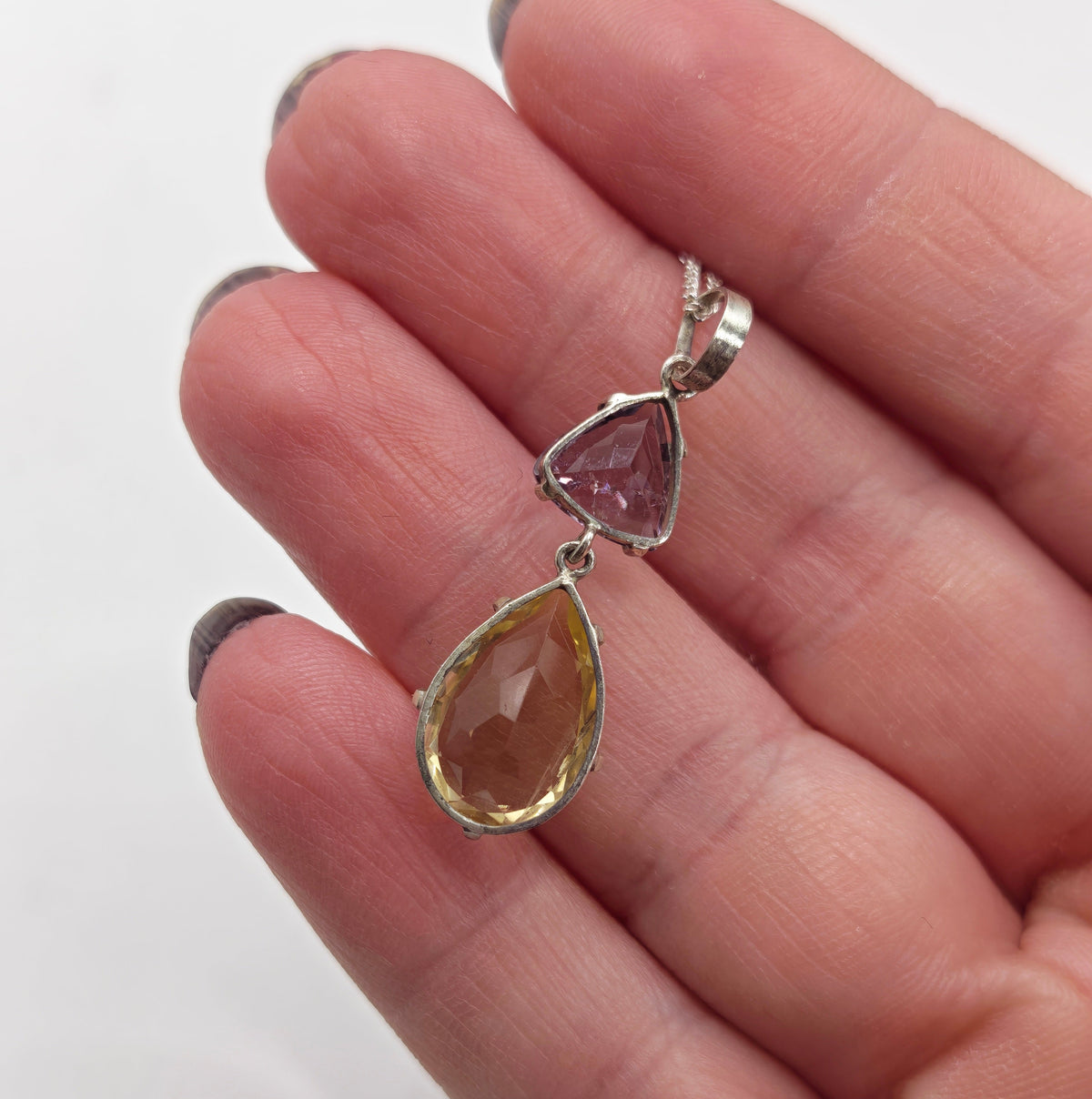 A silver pendant necklace featuring a larger citrine teardrop and a smaller triangular amethyst gemstone, im hand