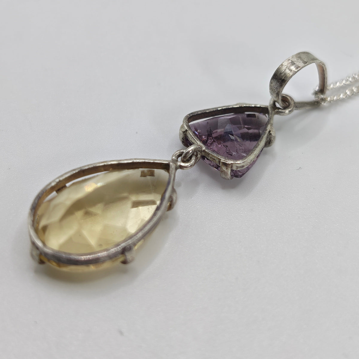 A silver pendant necklace featuring a larger citrine teardrop and a smaller triangular amethyst gemstone, shown on a plain light background.