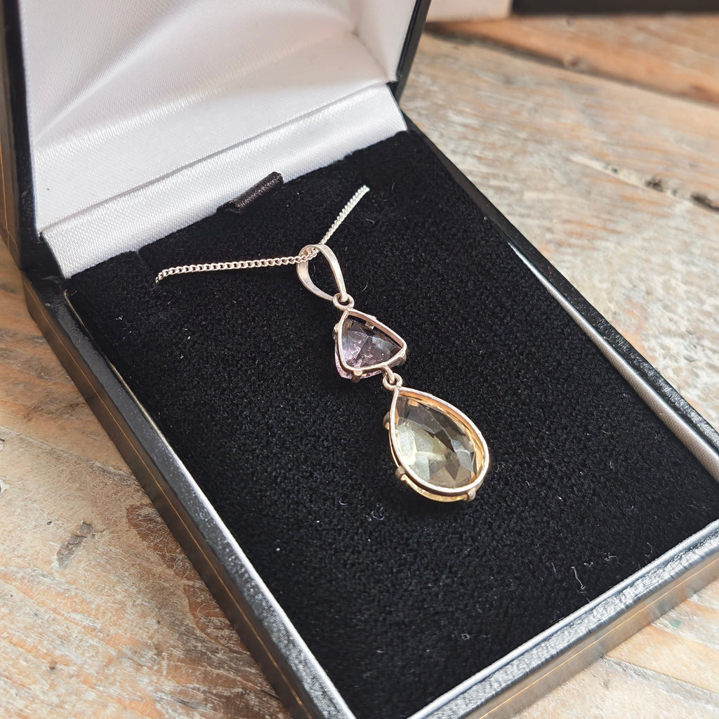 A silver pendant necklace featuring a larger citrine teardrop and a smaller triangular amethyst gemstone,on wooden rustic side closeup