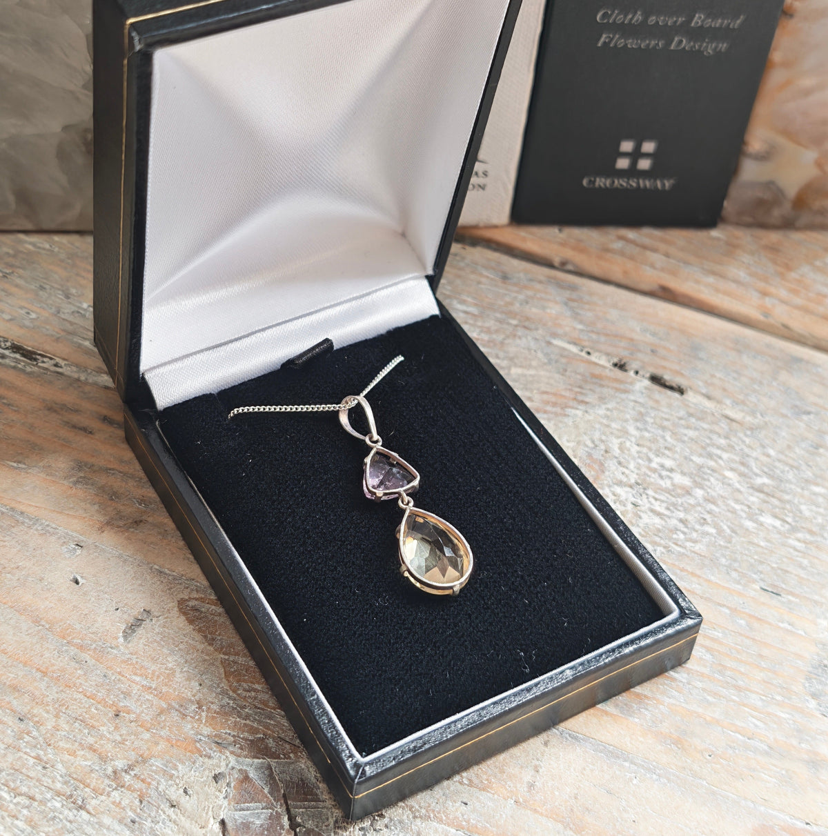 A silver pendant necklace featuring a larger citrine teardrop and a smaller triangular amethyst gemstone,on wooden rustic side