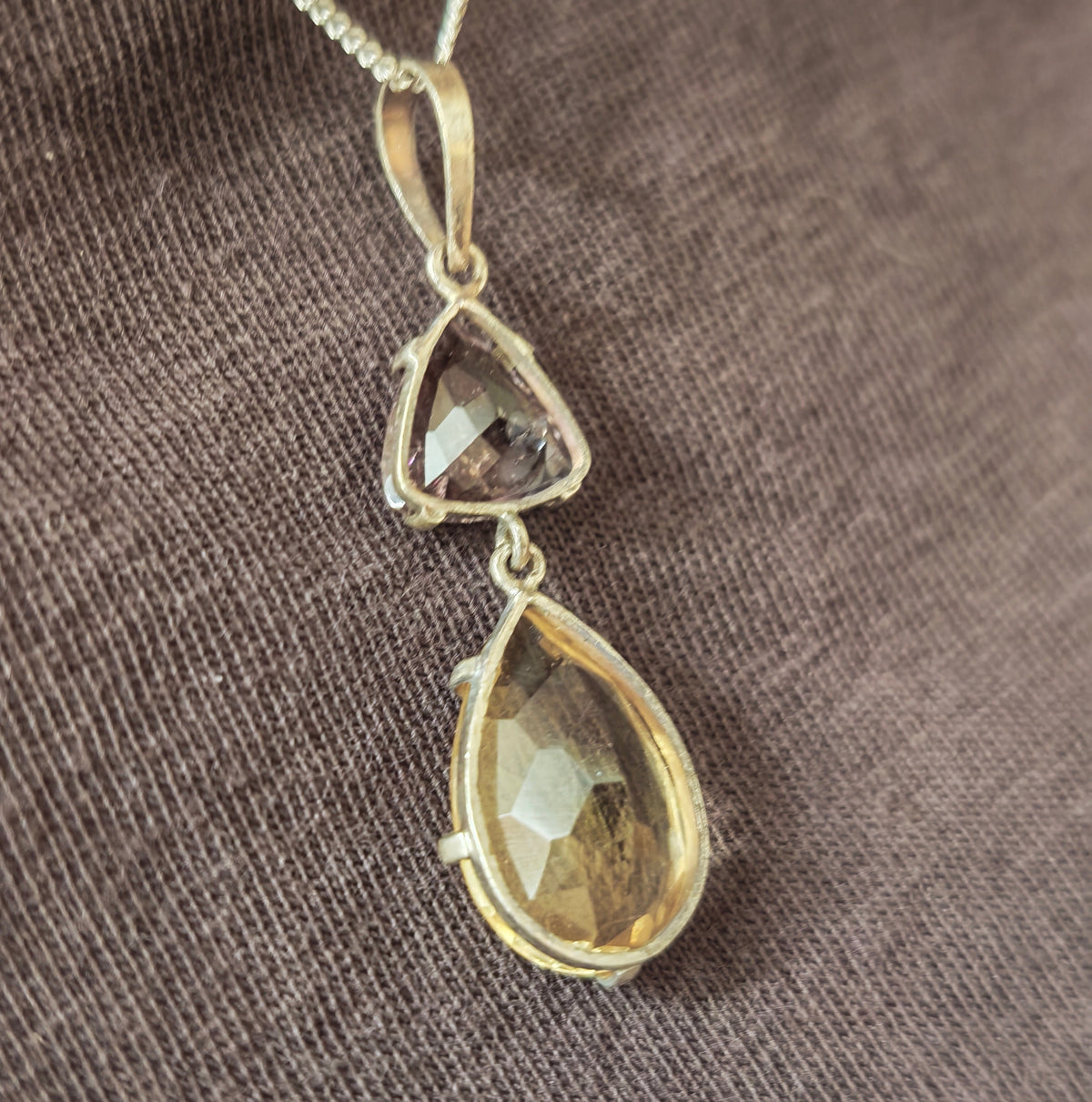 A silver pendant necklace featuring a larger citrine teardrop and a smaller triangular amethyst gemstone, being worn close up resting on chest