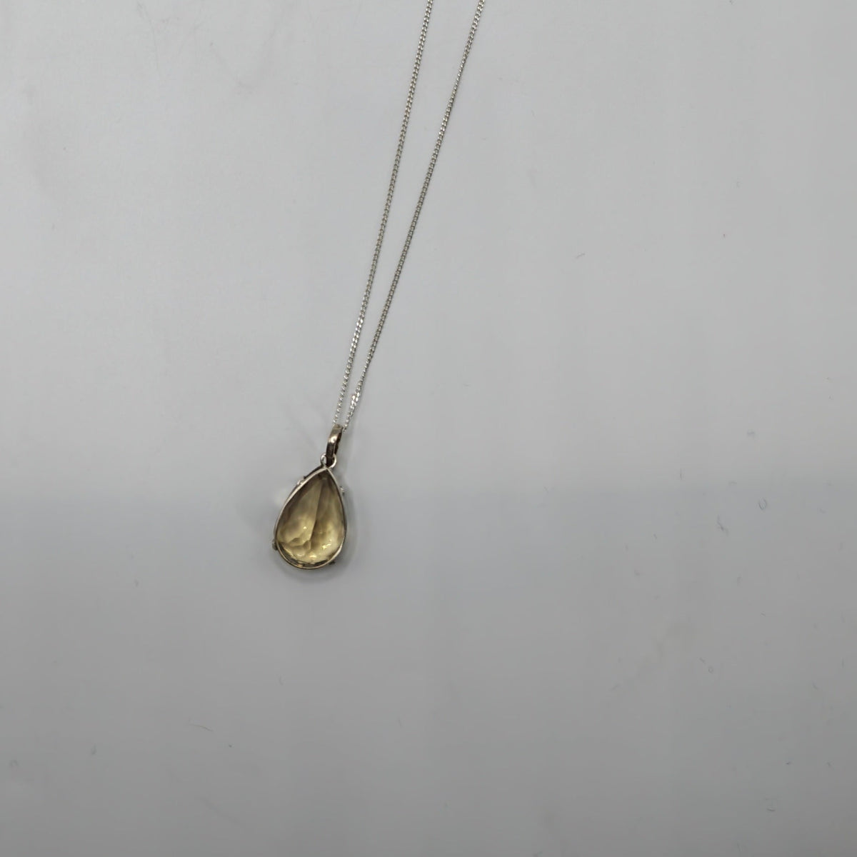 A delicate silver chain necklace with a small Citrine Faceted teardrop-shaped  gemstone pendant, displayed against a plain light  background.