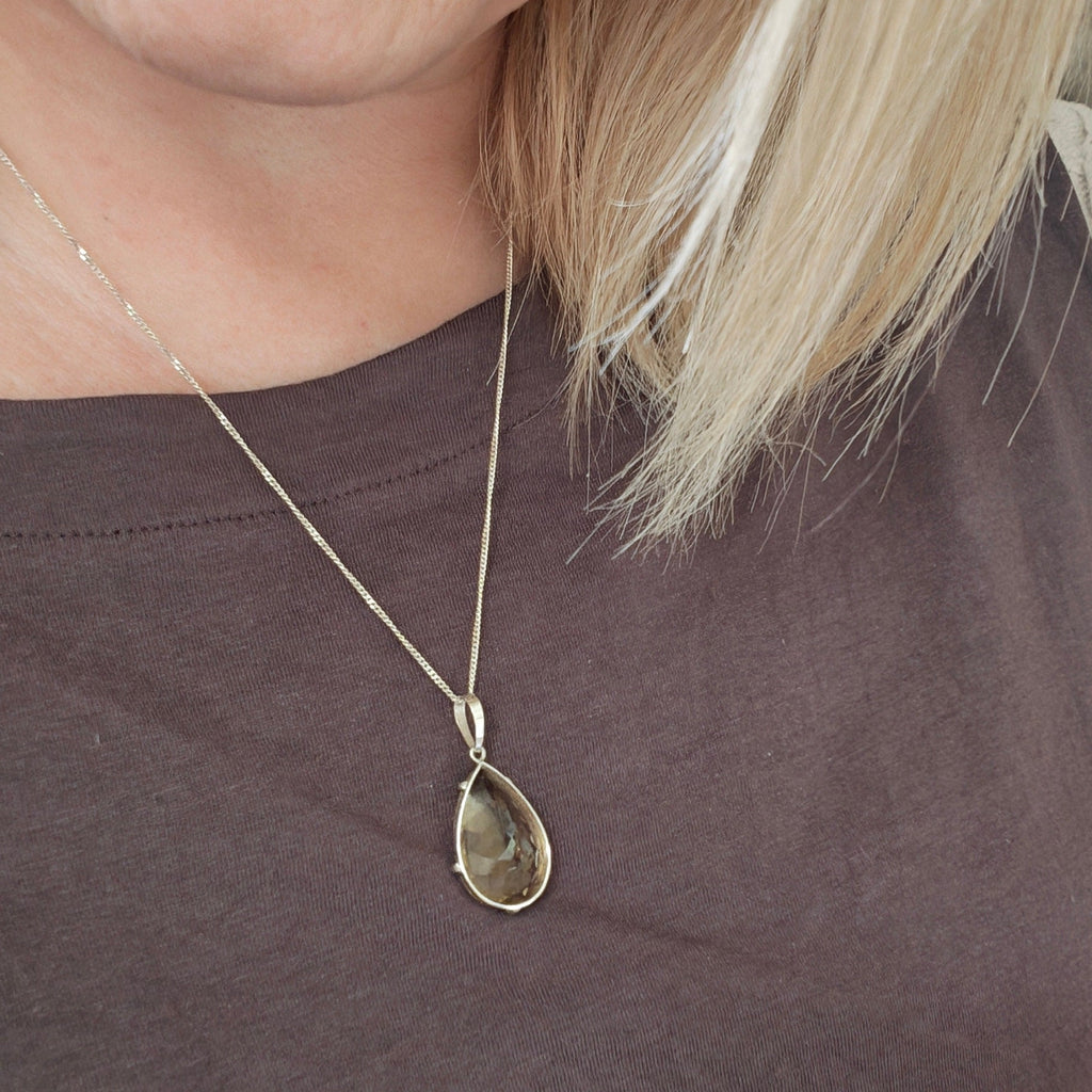 A close-up of a woman wearing a delicate hand faceted citrine pendant set in silver with a silve chain necklace, paired with a dark brown top.