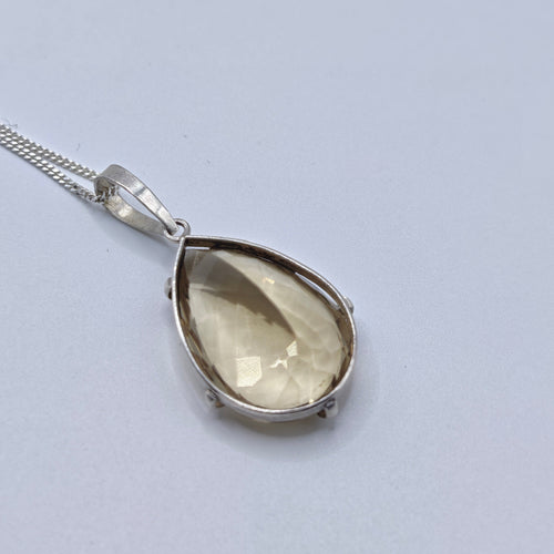 A teardrop-shaped pendant necklace featuring a faceted light yellow citrine gemstone in a silver bezel setting, suspended from a silver chain against a white background.