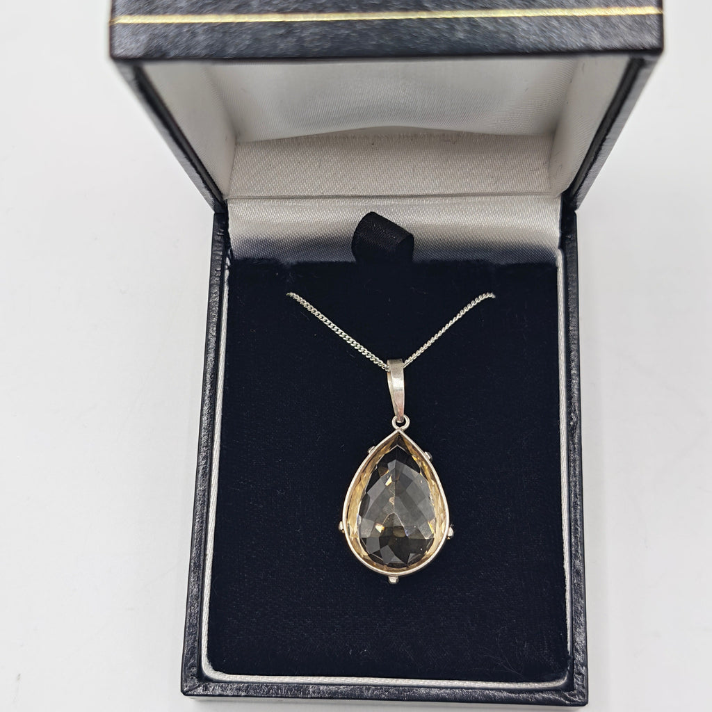A teardrop-shaped pendant necklace with a hand faceted citrine gemstone in a silver setting, displayed inside an open black jewelry box with a black velvet insert.