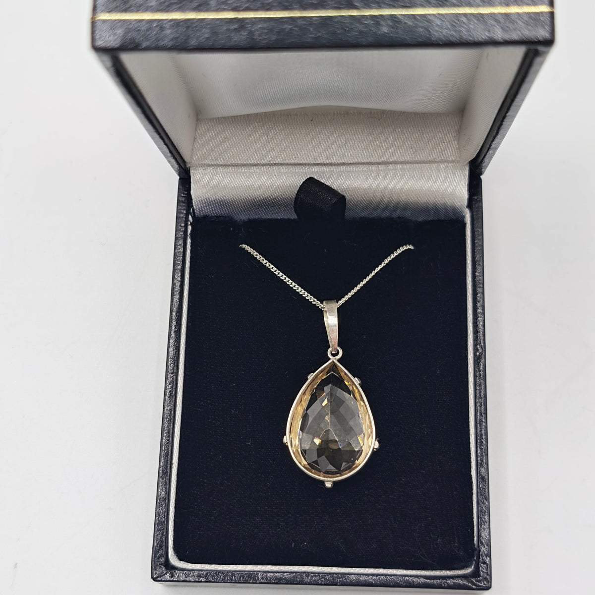 A teardrop-shaped pendant necklace with a hand faceted citrine gemstone in a silver setting, displayed inside an open black jewelry box with a black velvet insert.