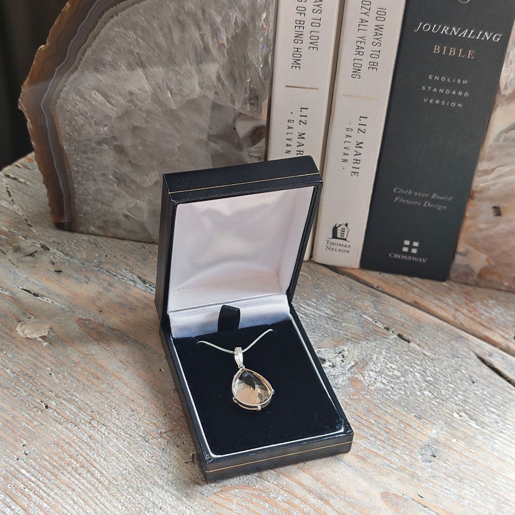 A Citrine teardrop pendant necklace displayed in an open black jewelry box with a white interior, set on a rustic wooden tabletop with books and an agate slice bookend in the background.