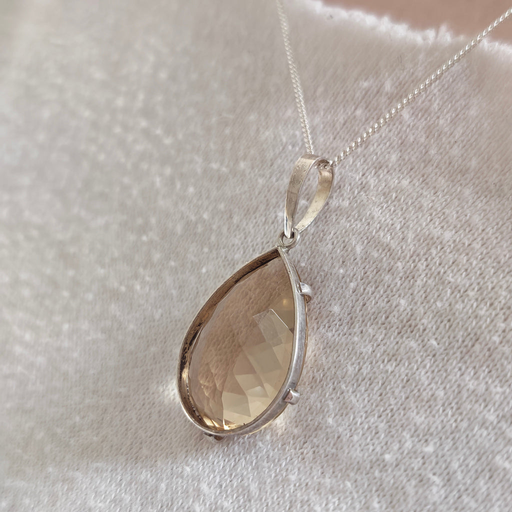 A  hand faceted teardrop-shaped champagne-colored citrine gemstone pendant in a silver bezel setting on a delicate silver chain, displayed on a textured white fabric background.