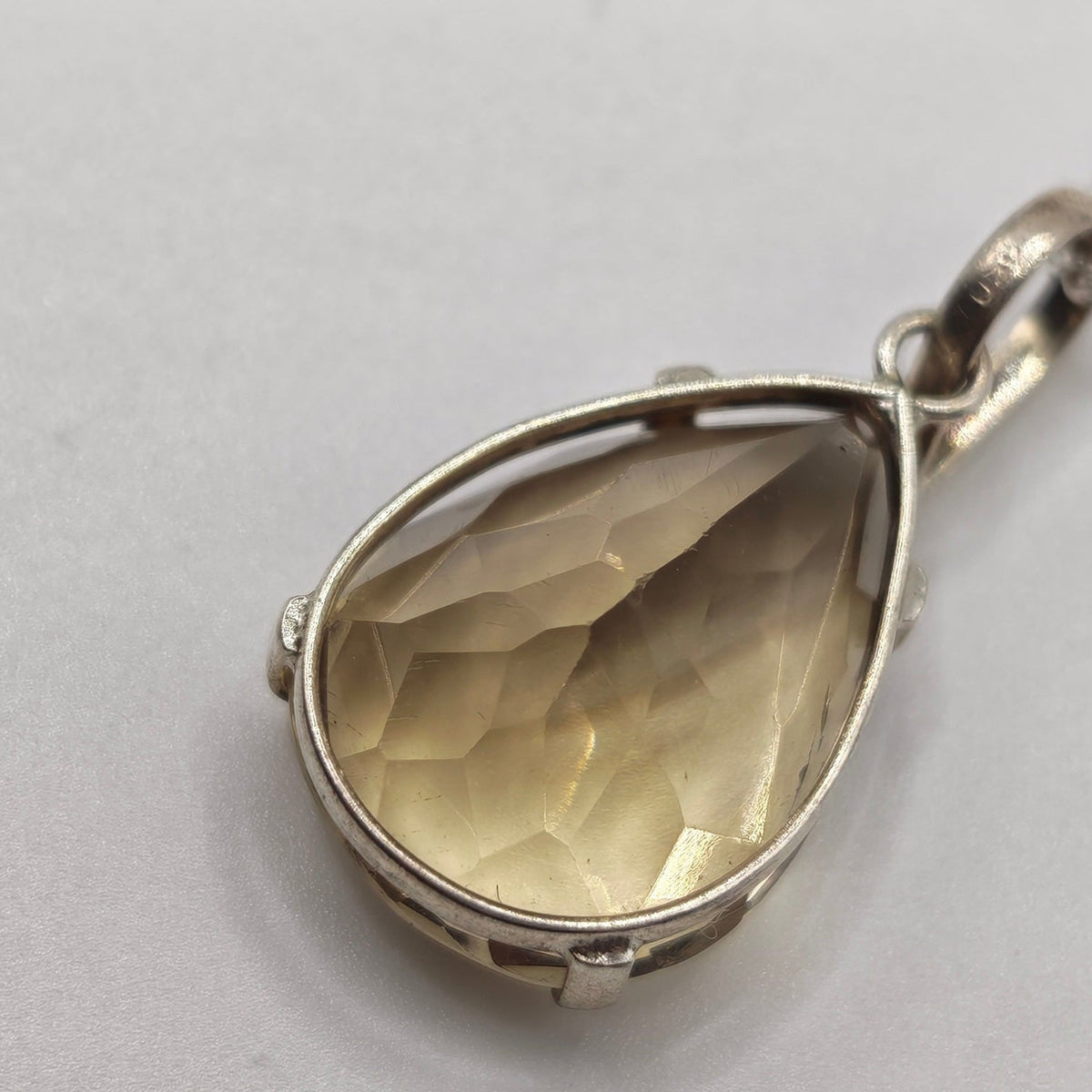 A teardrop-shaped pendant necklace featuring a faceted, Fasceted citrine gold-toned gemstone in a silver-toned bezel setting, suspended from a silver chain against a plain light gray background.