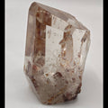 A raw, unpolished Fire quartz crystal with natural inclusions, standing upright on a neutral light gray background.