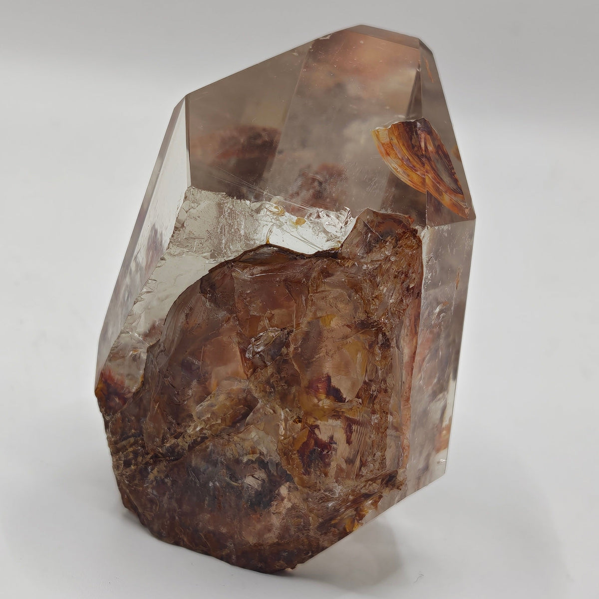 A faceted fire quartz freeform crystal specimen with a smoky translucent body and rich brown inclusions, photographed on a plain light gray background.