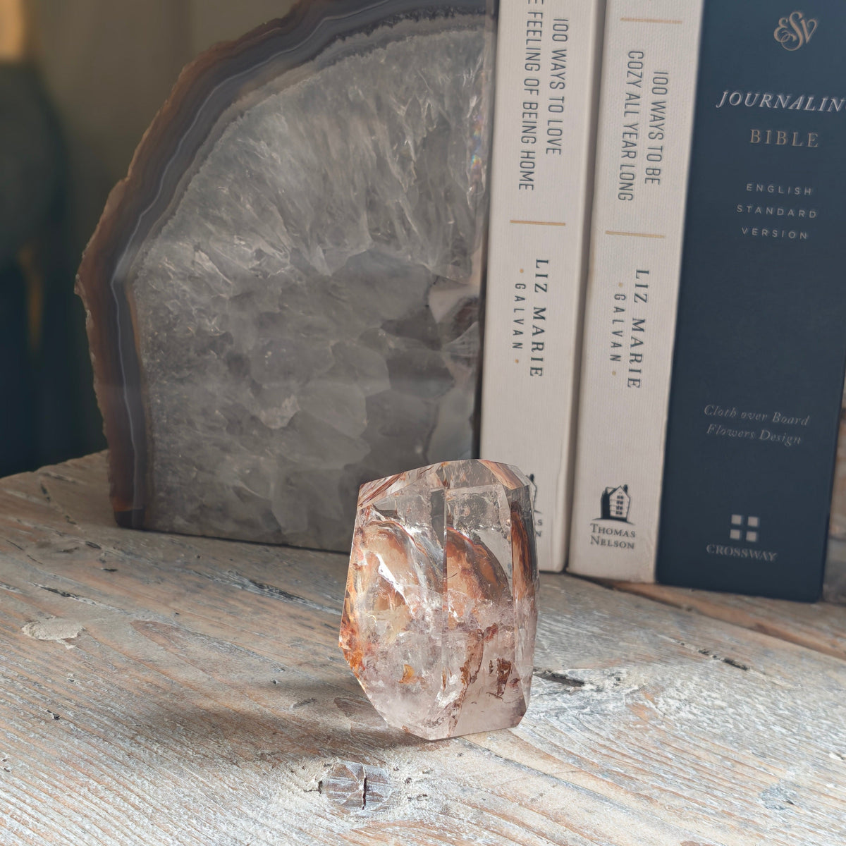 A raw, rough Fire quartz crystal sits on a distressed white wooden tabletop, styled with books and a large polished agate slice in the background.