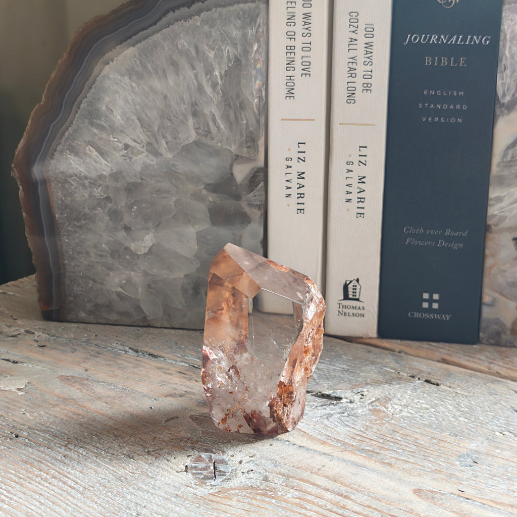 A raw, rough Fire quartz crystal from a side view sits on a distressed white wooden tabletop, styled with books and a large polished agate slice in the background