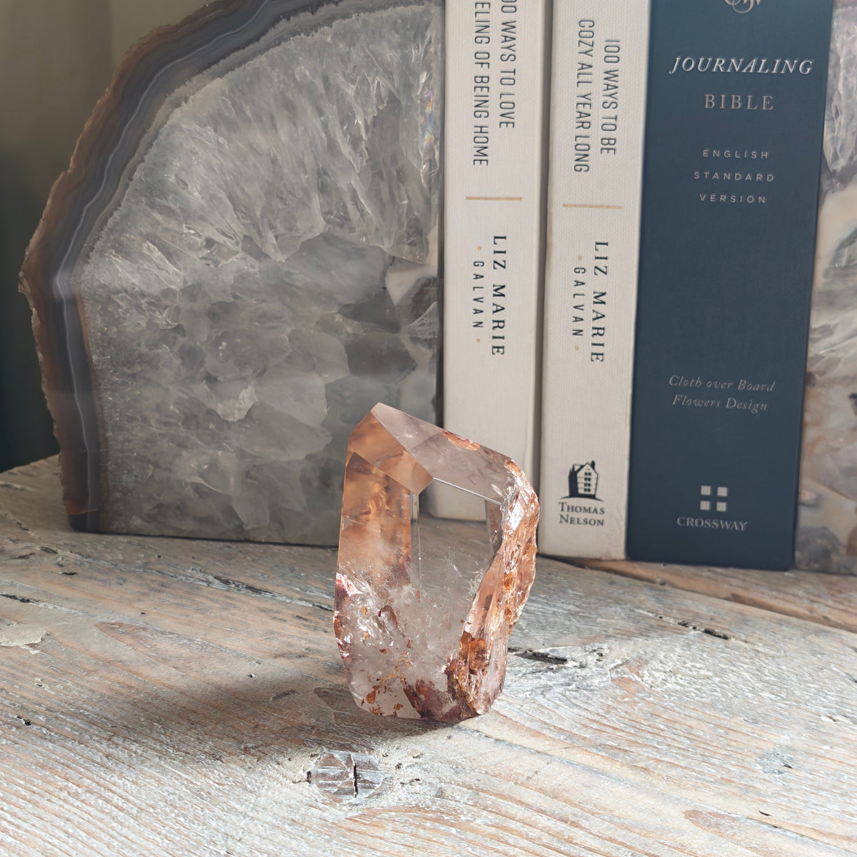 A raw, rough Fire quartz crystal from a side view sits on a distressed white wooden tabletop, styled with books and a large polished agate slice in the background
