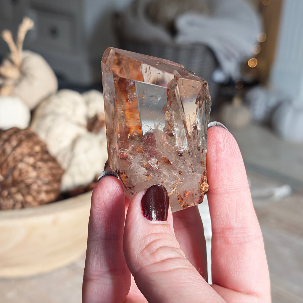 A hand with dark burgundy nail polish holds a raw, faceted fire quartz crystal with brown and amber inclusions, set against a softly blurred home decor background.