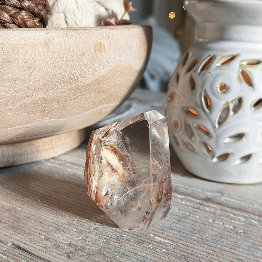 A faceted fire quartz crystal with natural hematite inclusions, standing on a rustic wooden table next to a white ceramic candle holder and a wooden bowl.