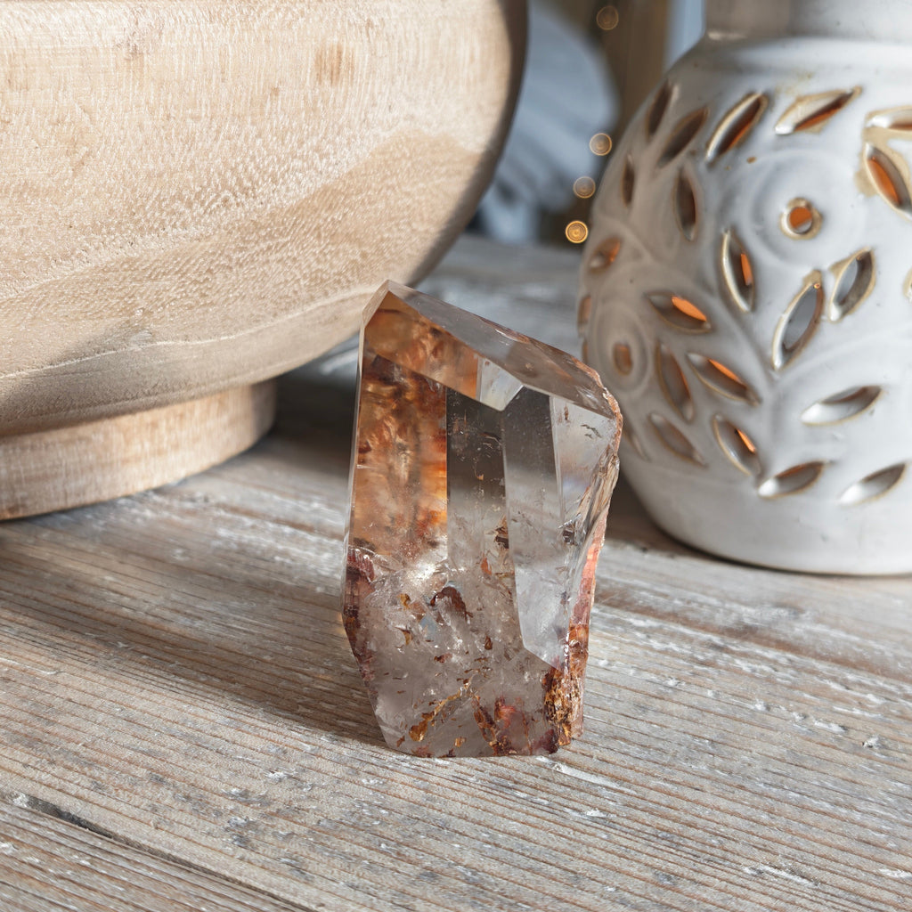 The opposite side of the faceted fire quartz crystal with natural hematite inclusions, standing on a rustic wooden table next to a white ceramic candle holder and a wooden bowl.