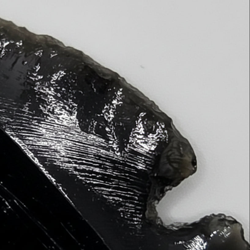 Close up of black obsidian arrow head front face showing hand-shaped edges, glossy black surface, and natural conchoidal fracture pattern