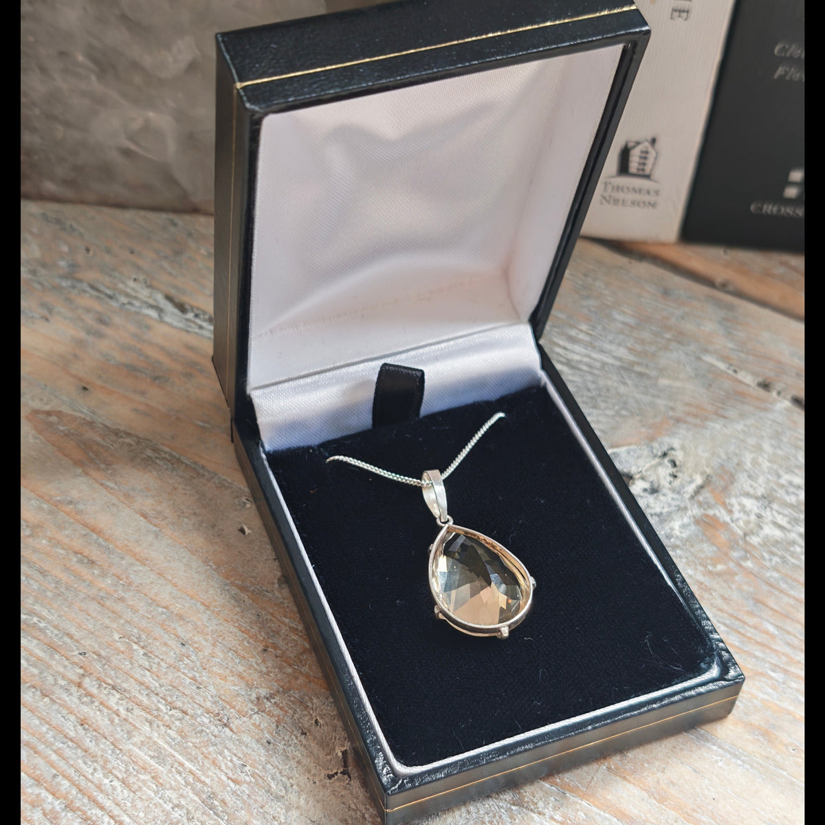 A silver teardrop pendant necklace with a hand faceted citrine gemstone, displayed inside an open black jewelry box with a black velvet lining, set on a rustic wooden table.