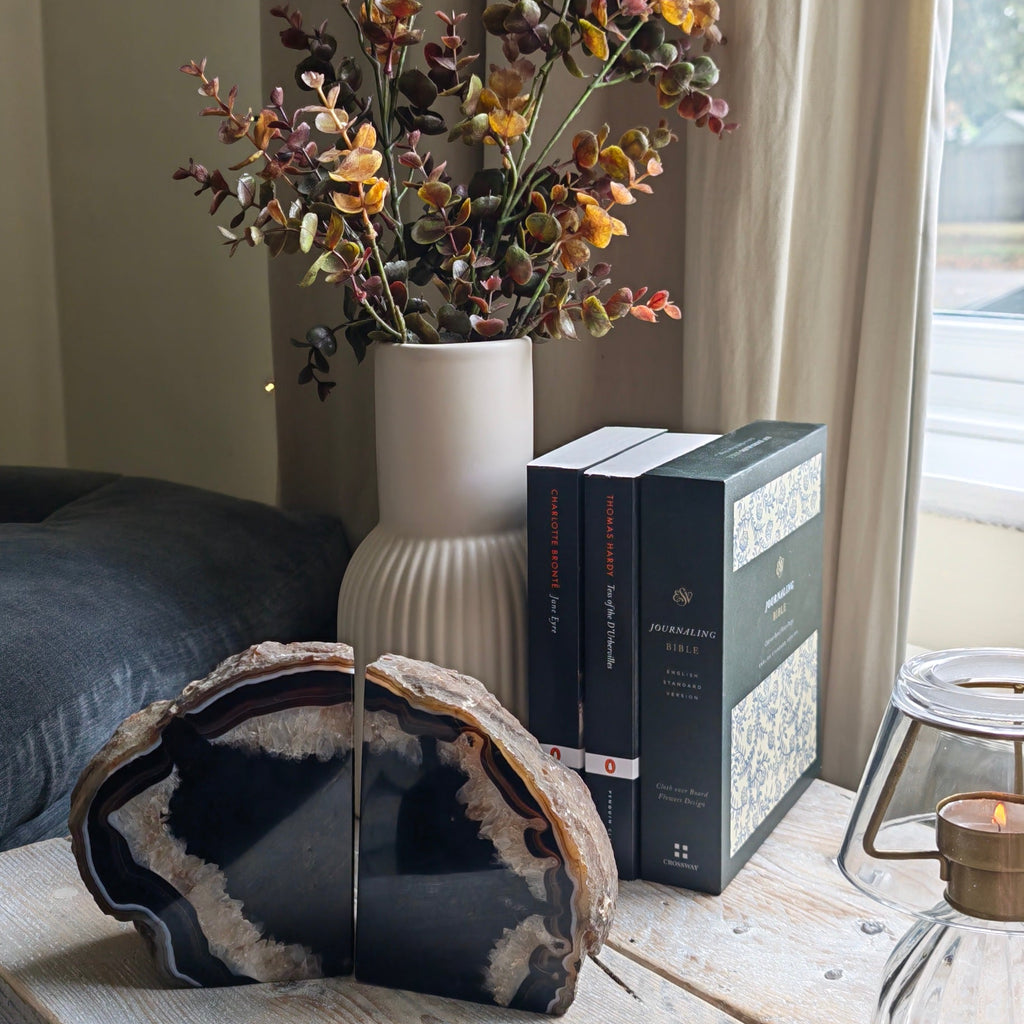 A split black and white agate stone bookends on a rustic white wooden table next to a ceramic vase with dried autumn flowers and a stack of books.