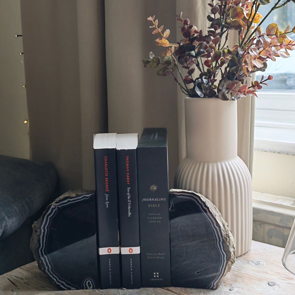 A pair of black Agate bookends with white banding holding three books on a rustic wooden table, next to a beige ribbed vase with dried flowers in a home setting.