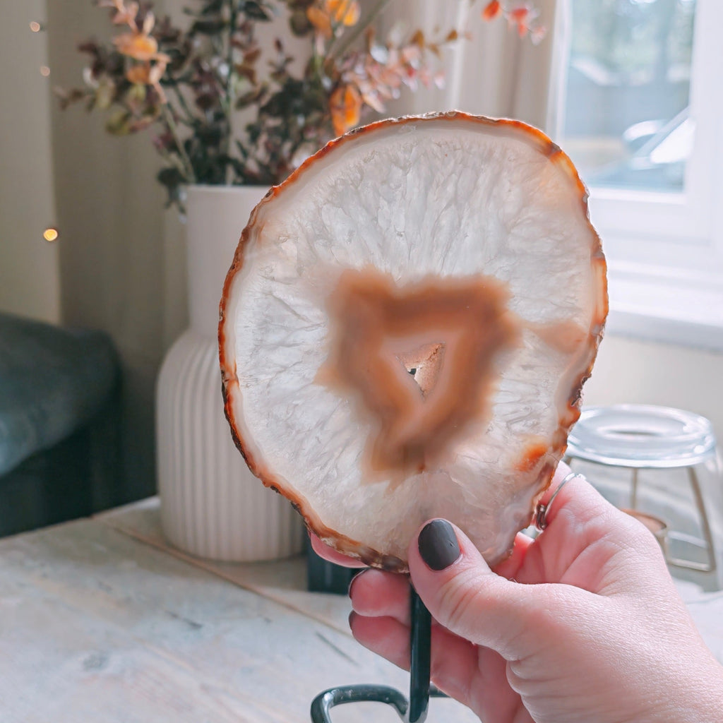 A hand with black nail polish holds a polished agate slice on a small black metal stand. The background shows a rustic white wooden table, a vase with dried flowers, and a window with soft natural light.