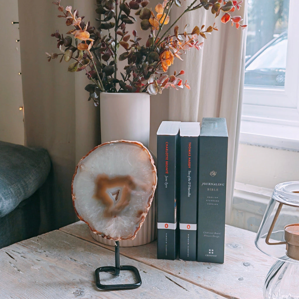 A rustic white wooden table styled in a cozy room setting, featuring a decorative agate slice on a stand, a vase of dried orange and brown flowers, and a stack of three dark gray books.