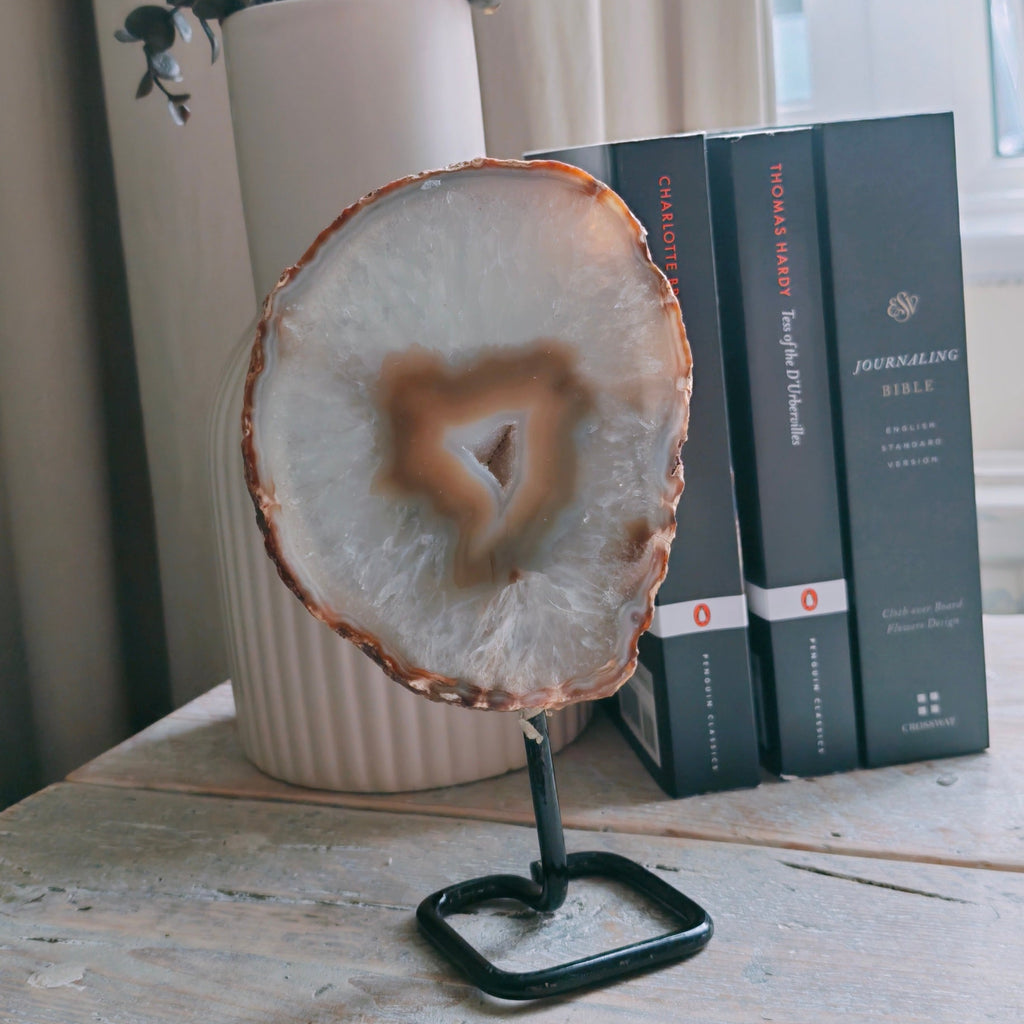 A polished agate slice with a druzzy center on a black metal stand, displayed on a light wood table next to books and a vase with dried flowers.
