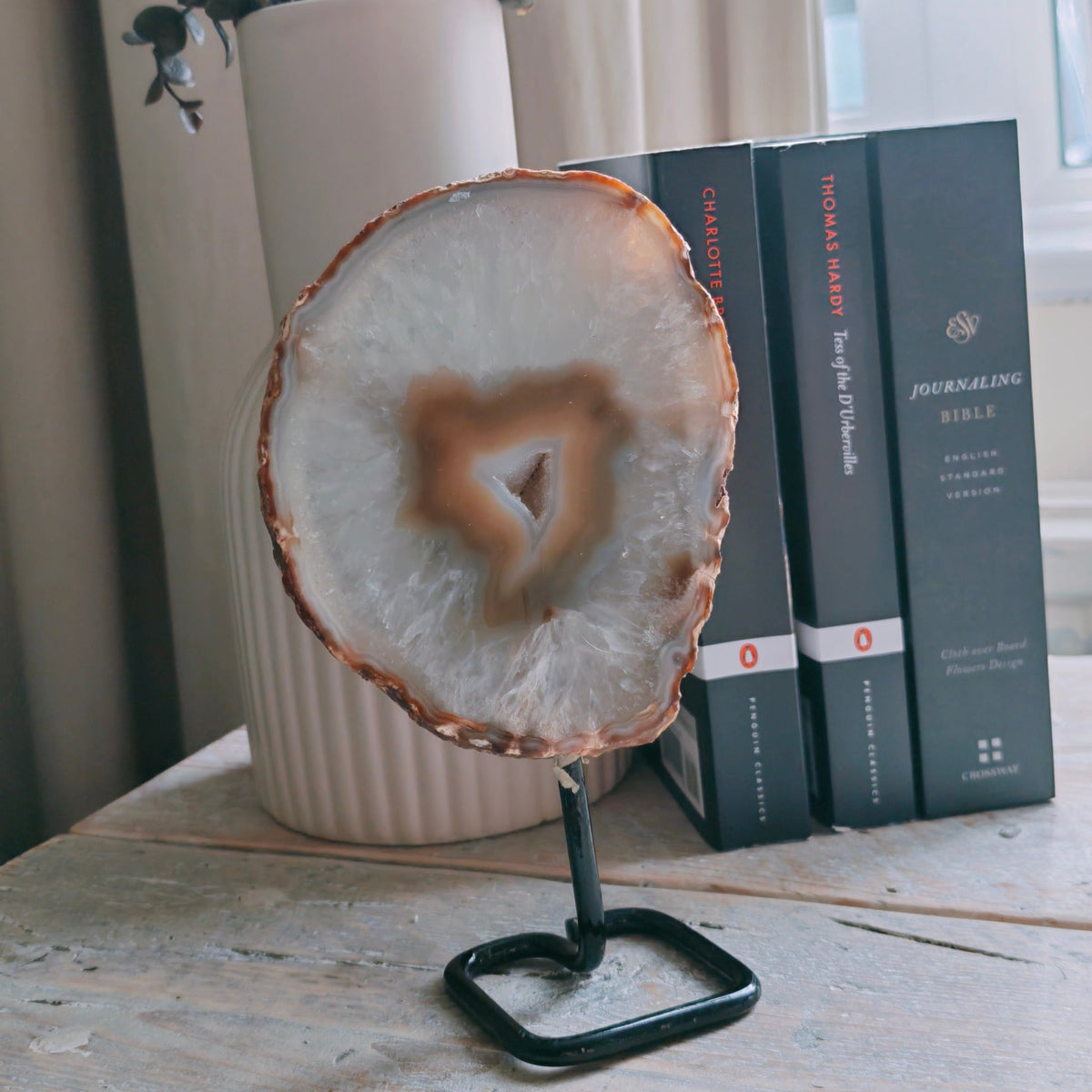 A polished agate slice with a druzzy center on a black metal stand, displayed on a light wood table next to books and a vase with dried flowers.