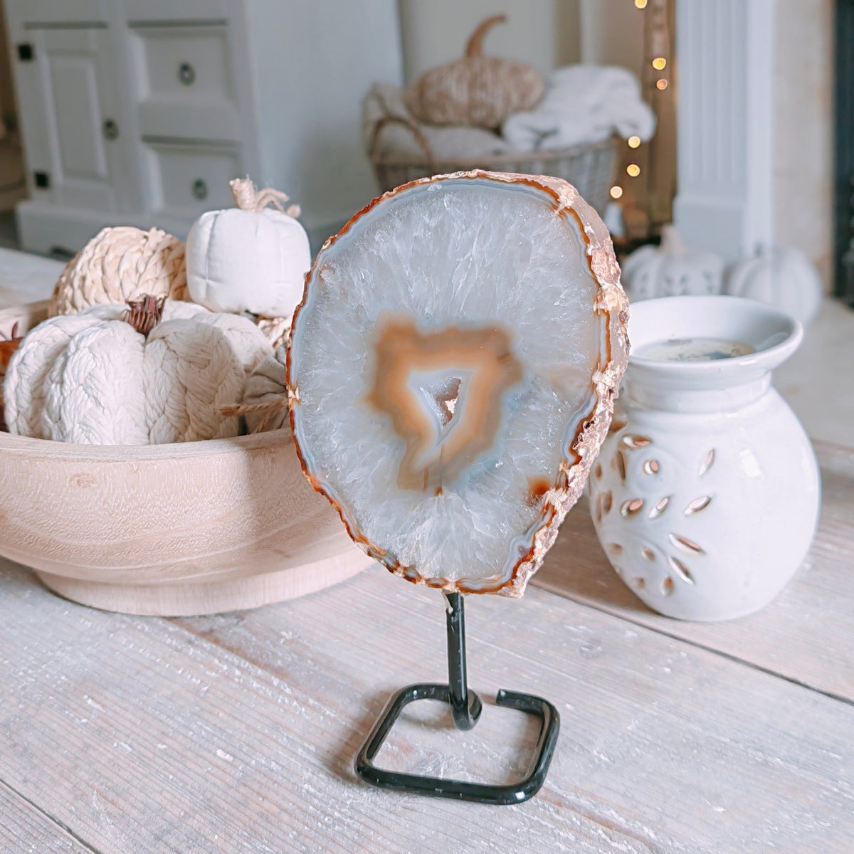 A round agate slice stand with a natural wood edge and a black metal base, displayed on a rustic white wooden table in a cozy, softly lit room with pumpkins and a fireplace in the background.