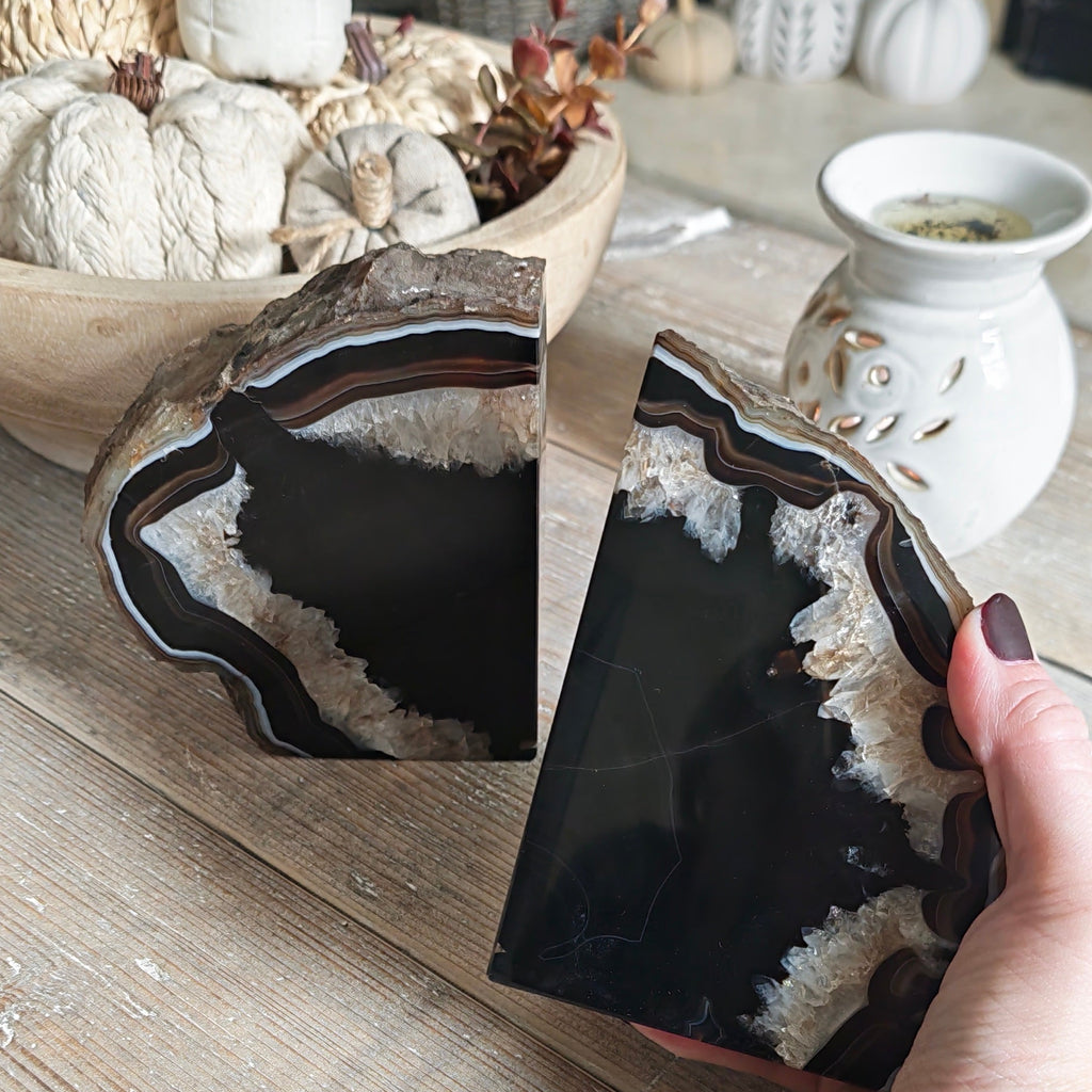 A person holds one half of a polished black agate geode slice on a rustic wooden table, with the other half resting on the table. The setting is a cozy, decorated room with autumn-themed decor like pumpkins.