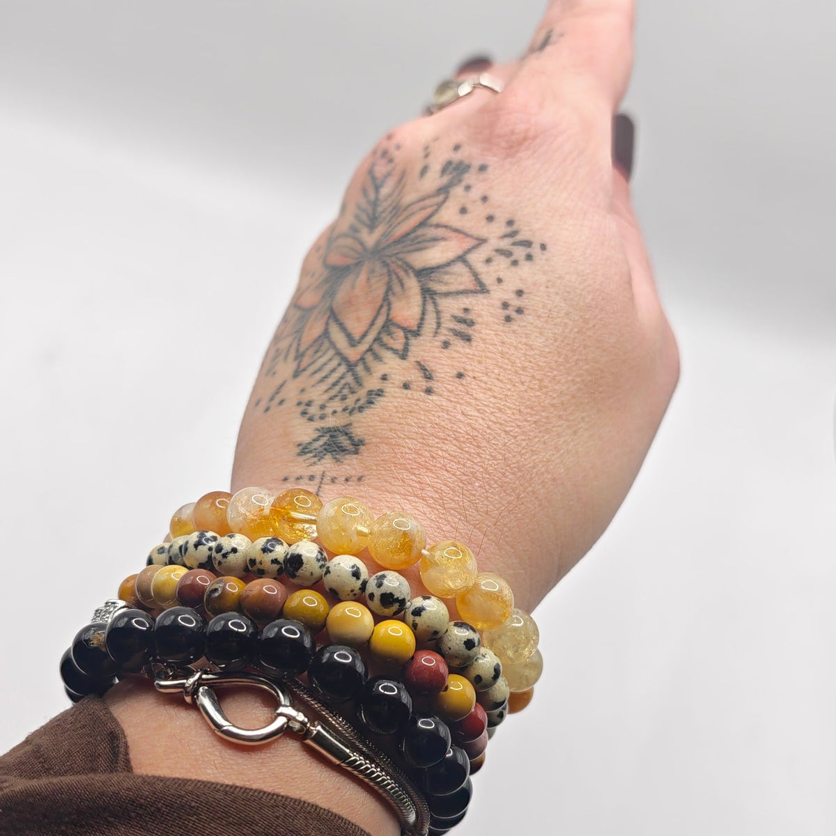 A close-up of a person's hand wearing multiple beaded bracelets, including yellow, black, and brown beads, with a floral tattoo on the back of the hand.
