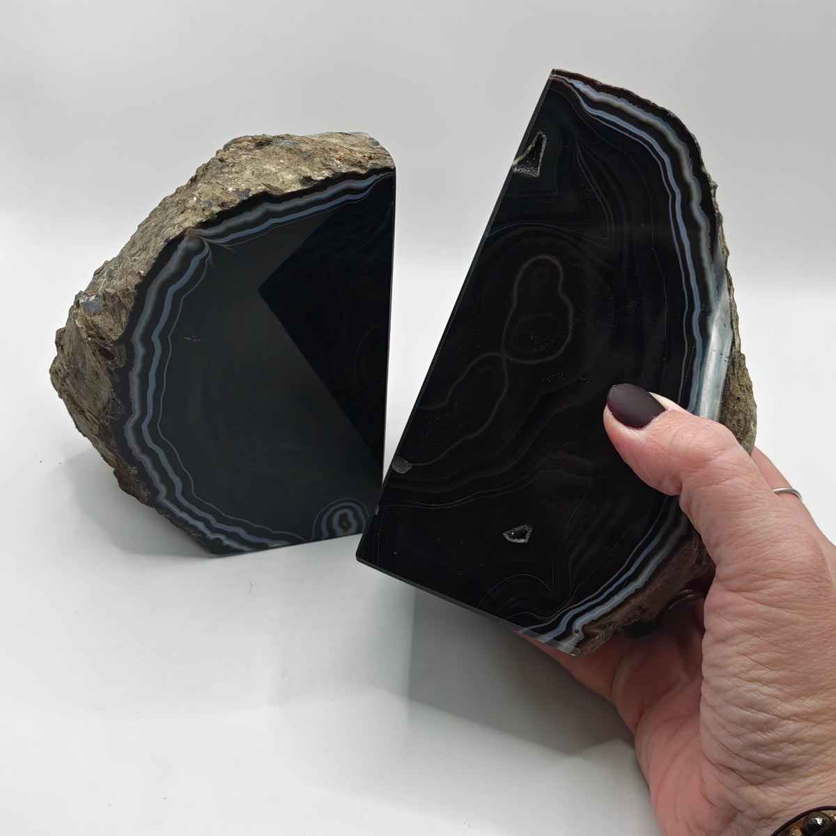 Two polished black agate geode halves with natural brown matrix and white-gray banded edges, displayed on a white surface with a hand holding one piece.