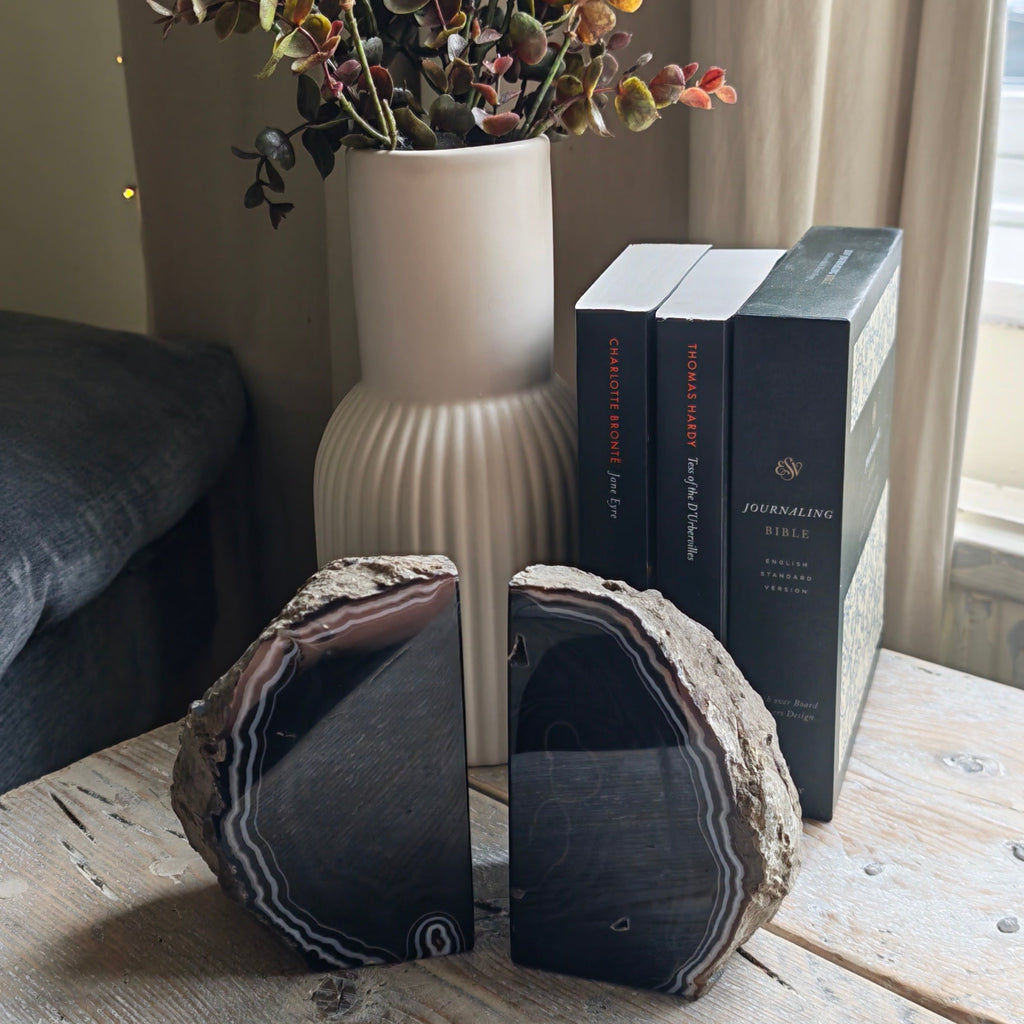 Two large agate geode bookends displayed on a rustic wooden table next to a white ribbed ceramic vase filled with dried flowers and a stack of books.