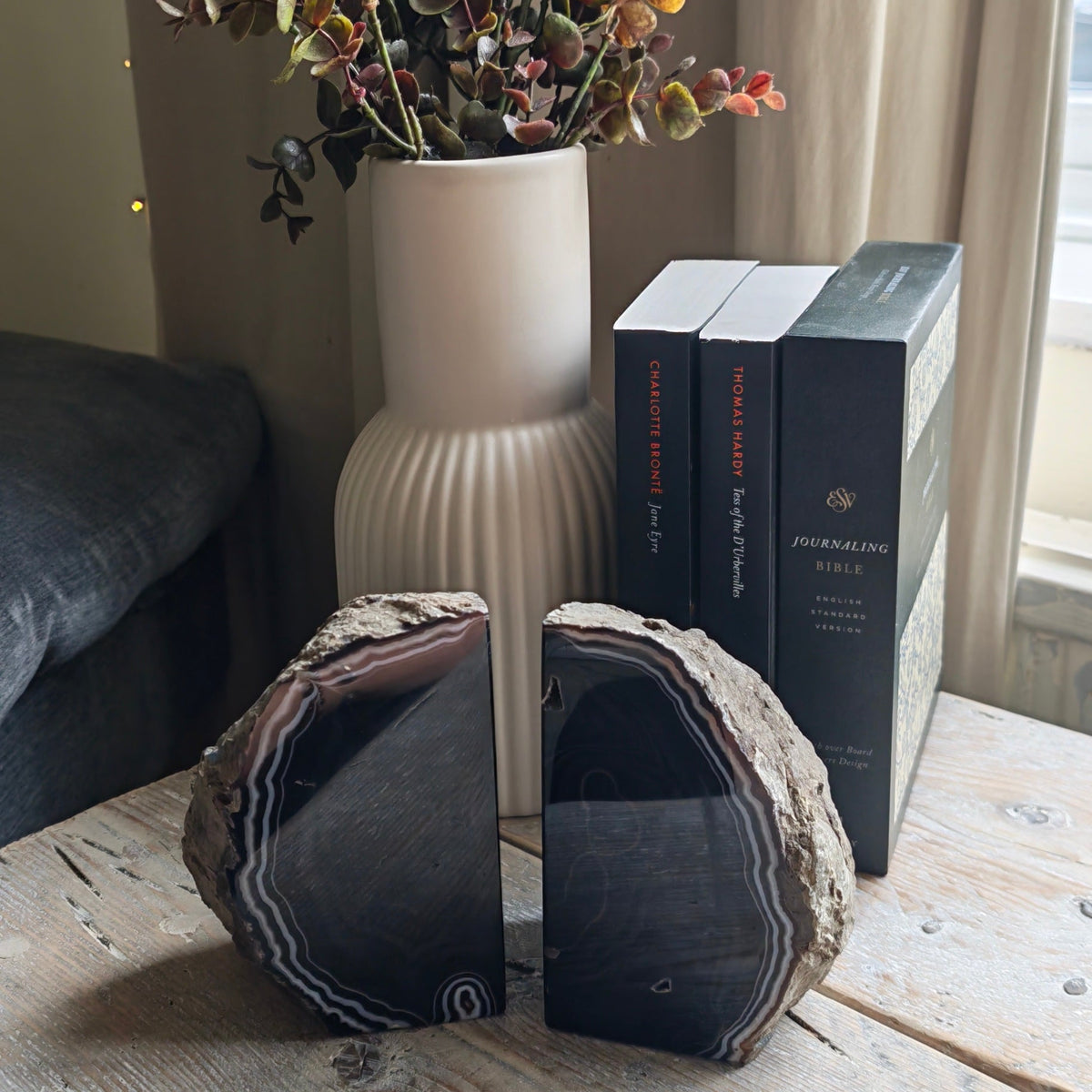 Two large agate geode bookends displayed on a rustic wooden table next to a white ribbed ceramic vase filled with dried flowers and a stack of books.