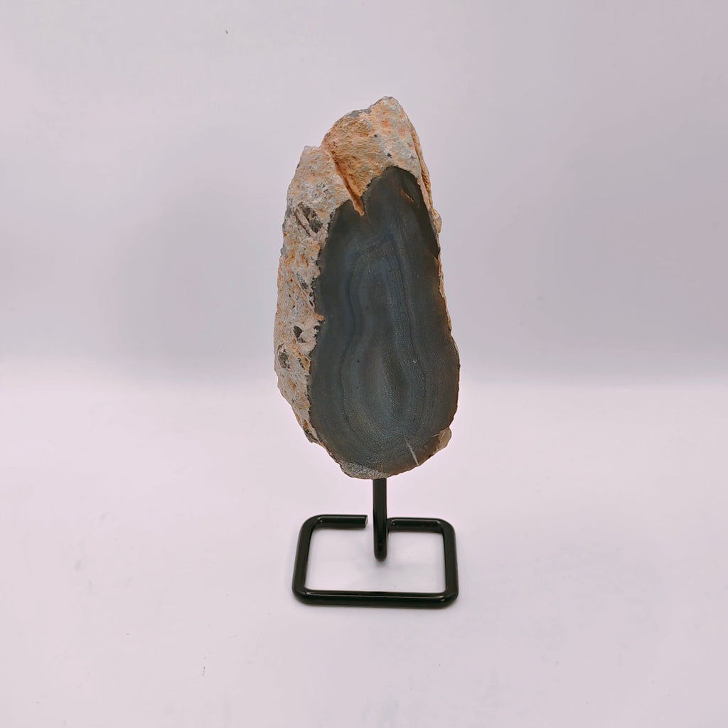 A polished geode slice with a dark gray interior and light brown outer crystalline skin, mounted on a small black metal display stand against a plain white background.
