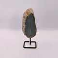 A polished geode slice with a dark gray interior and light brown outer crystalline skin, mounted on a small black metal display stand against a plain white background.