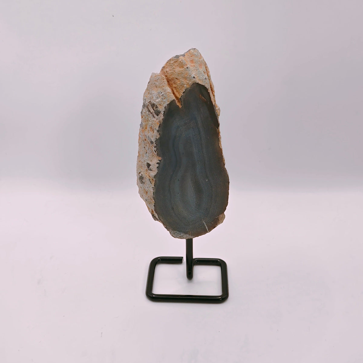 A polished geode slice with a dark gray interior and light brown outer crystalline skin, mounted on a small black metal display stand against a plain white background.