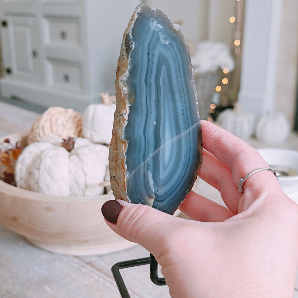 A hand holding a polished blue agate geode slice on a black display stand, set against a softly blurred home decor background with a white dresser and warm string lights.