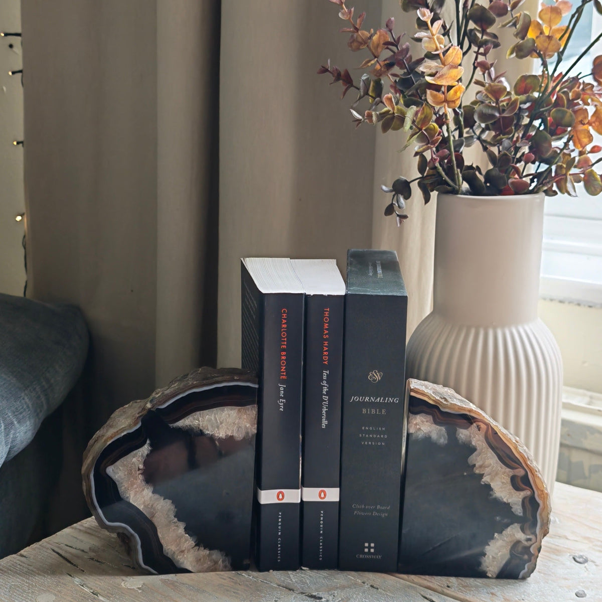 A pair of black agate stone bookends holding up three black books on a rustic wooden table, with a white ribbed ceramic vase of dried autumn flowers in the background near a window.